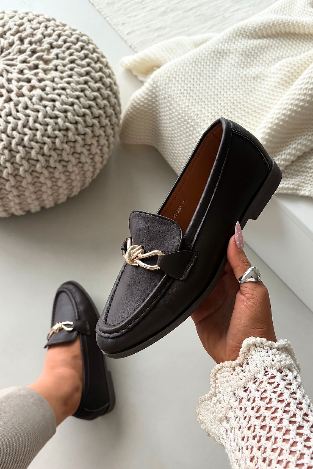 Black loafers with gold accents held by a hand against a neutral background