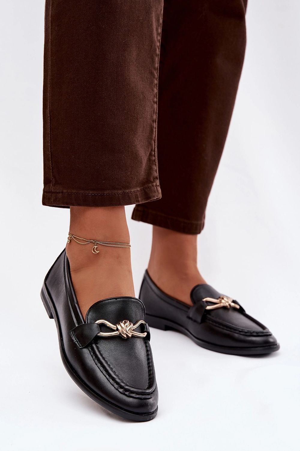 Black loafers with gold accents worn with brown pants on a white background