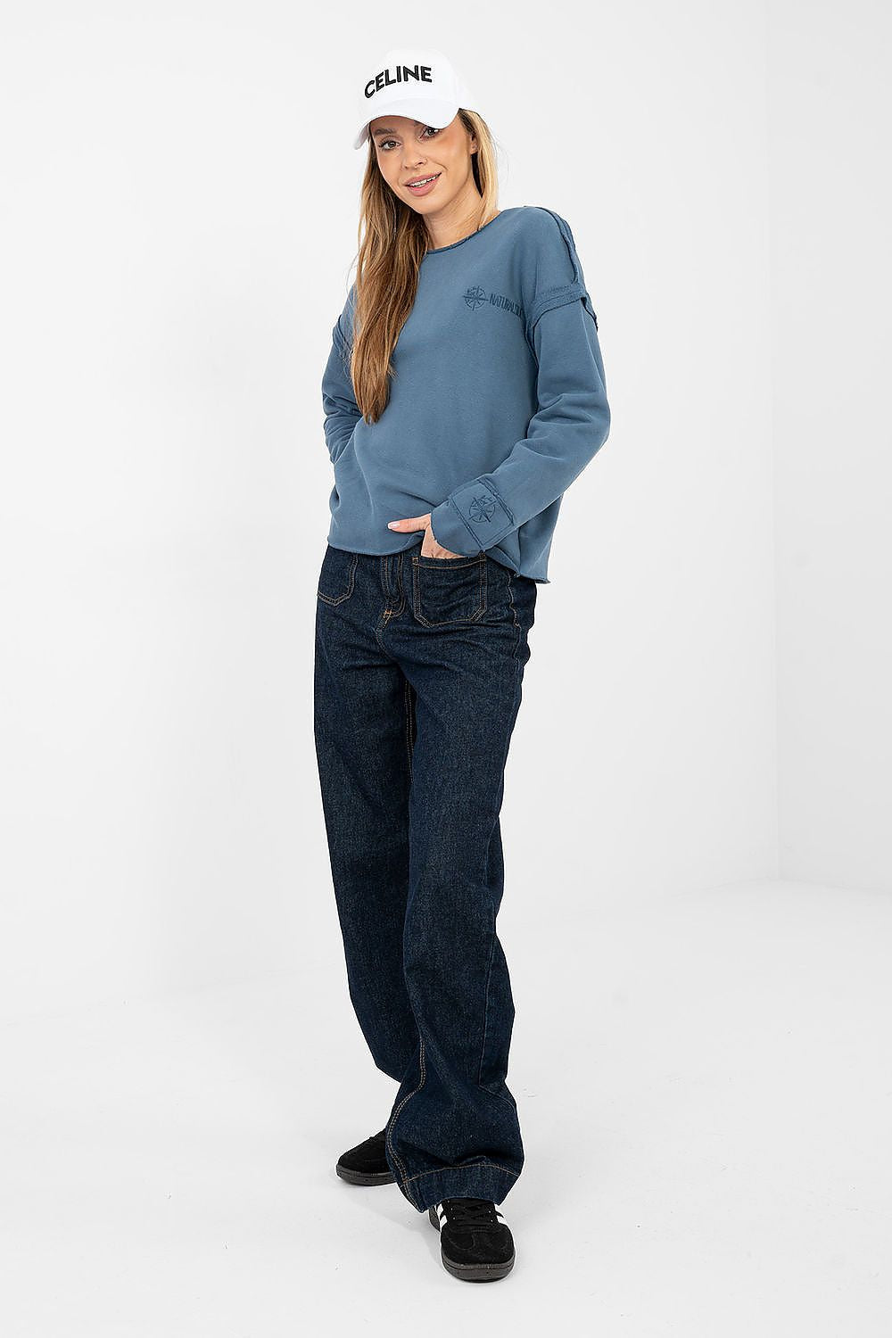 Woman wearing a blue sweatshirt, dark jeans, and a white cap with 'Celine' branding on a white background