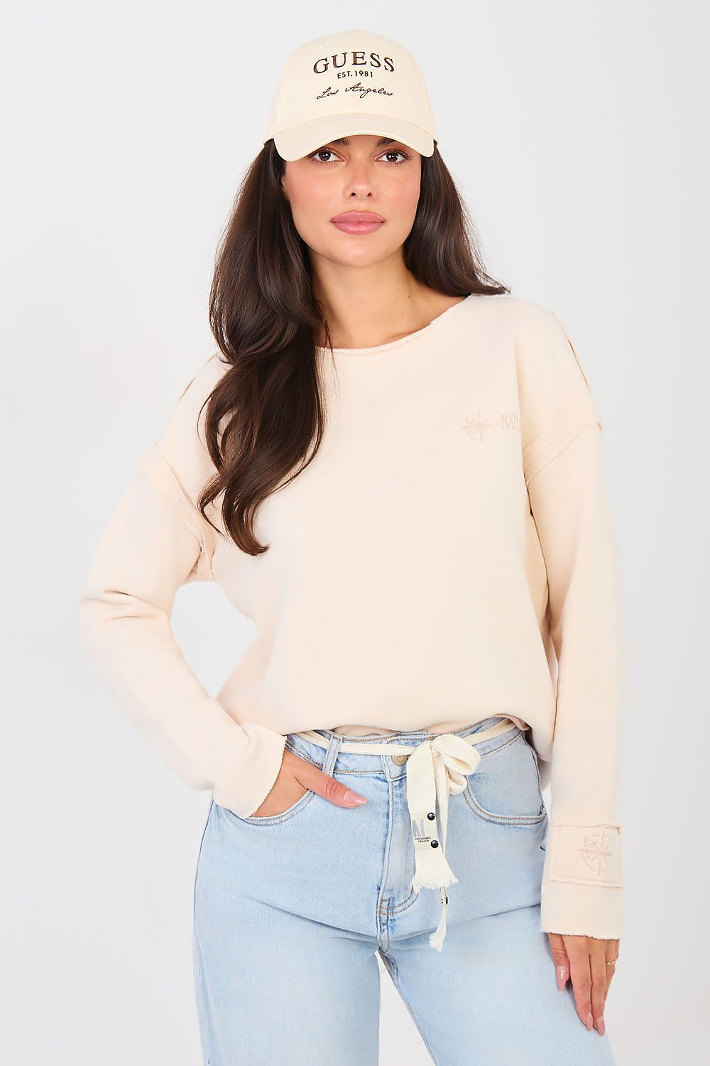 Woman wearing a beige sweater and light blue jeans with a beige cap on a white background