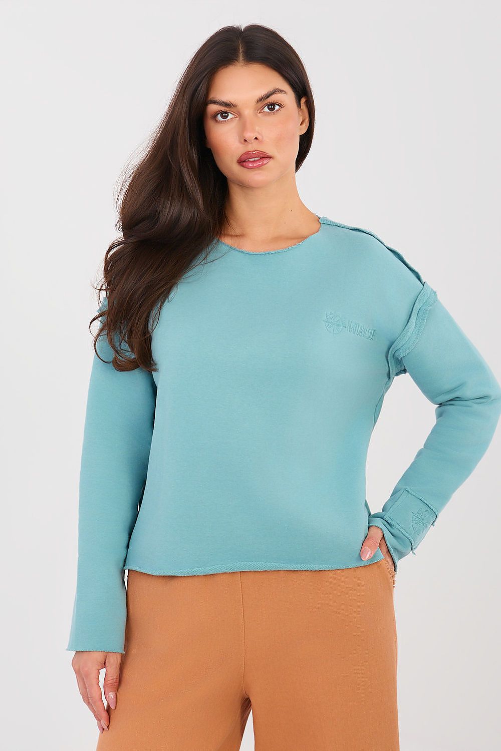 Woman wearing a teal sweater and brown pants against a white background