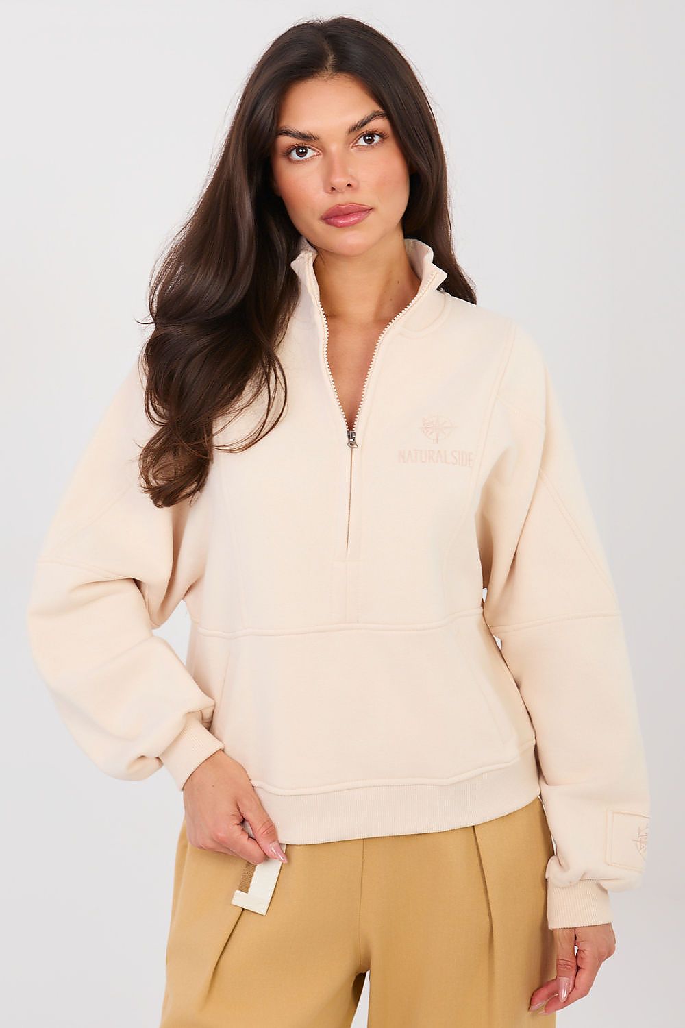 Woman wearing a beige half-zip sweatshirt and tan pants against a white background