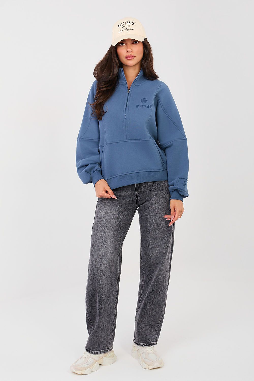 Person wearing a blue hoodie and gray jeans with a white cap on a light gray background
