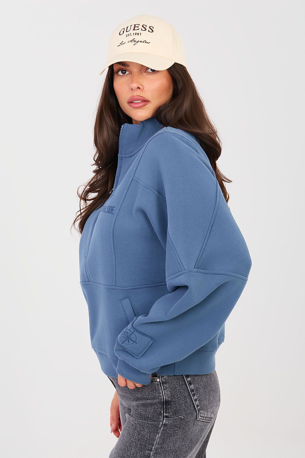 Woman wearing a blue sweatshirt and beige cap with 'Guess' logo on a white background