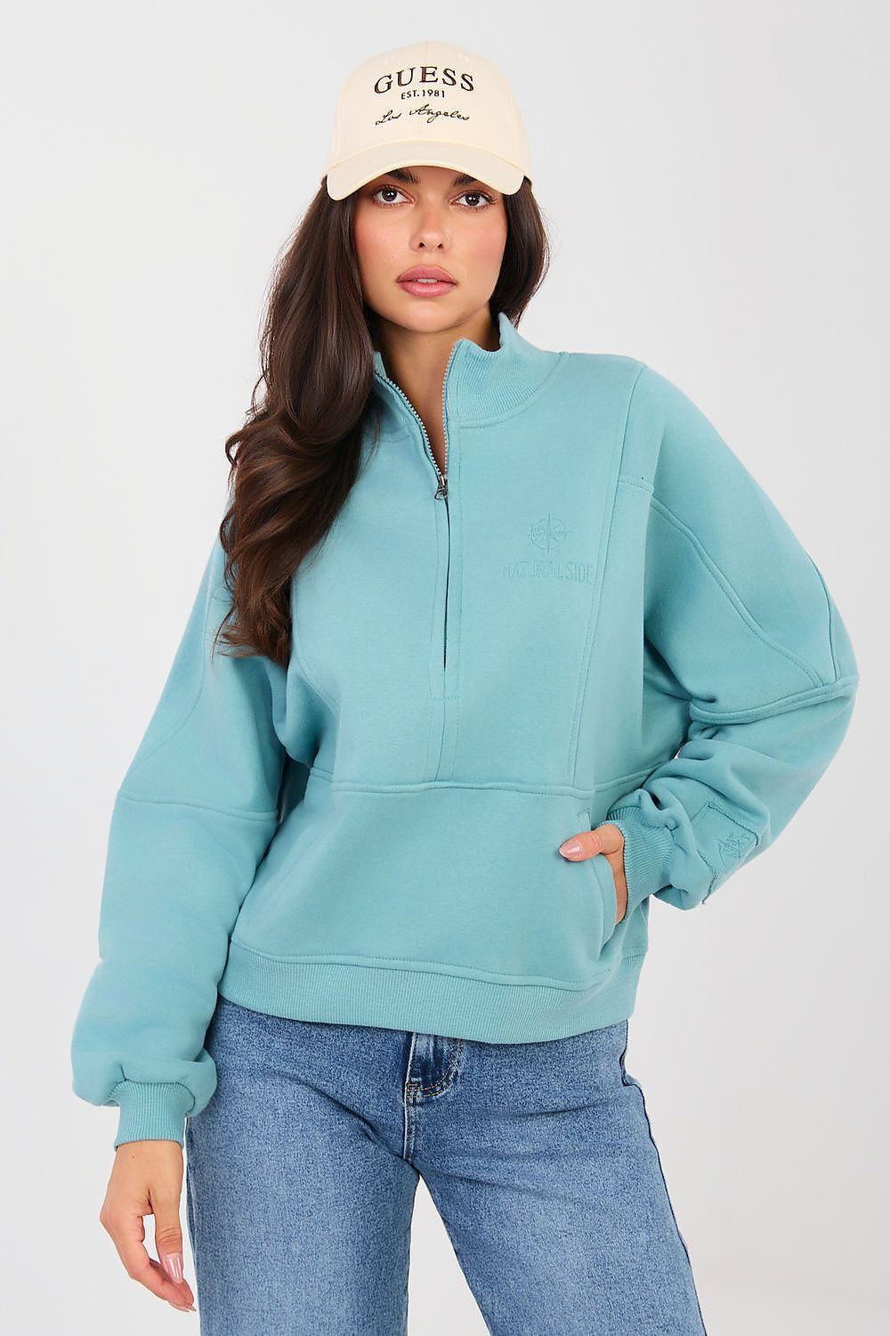 Woman wearing a teal half-zip sweatshirt and blue jeans with a beige cap on a white background