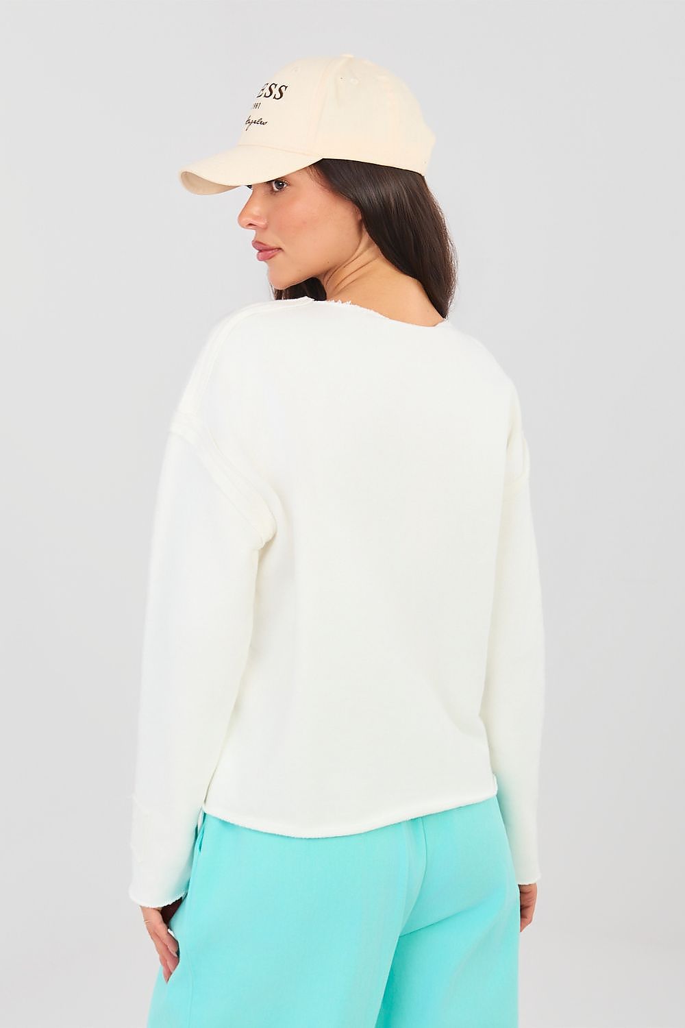 Woman wearing a white sweater and light blue pants with a beige cap on a plain background