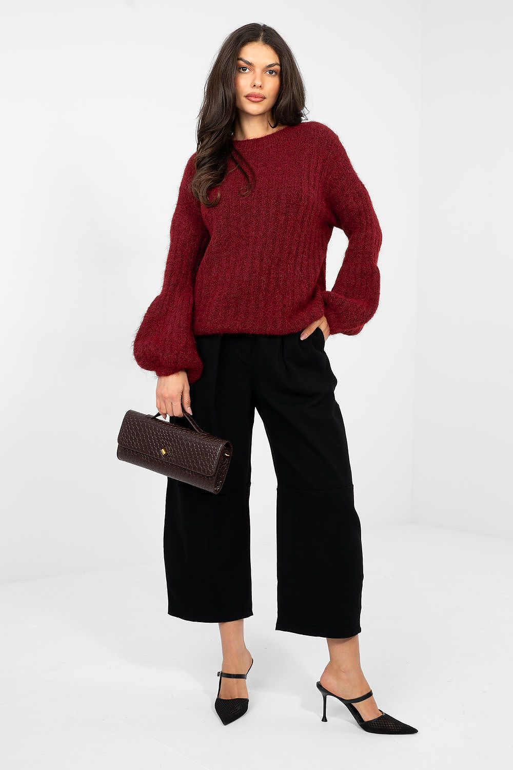 Woman wearing a red sweater and black pants holding a brown handbag on a white background