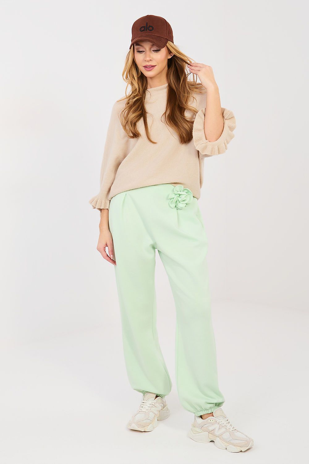 Woman wearing a beige sweater, green pants, and a brown cap on a white background