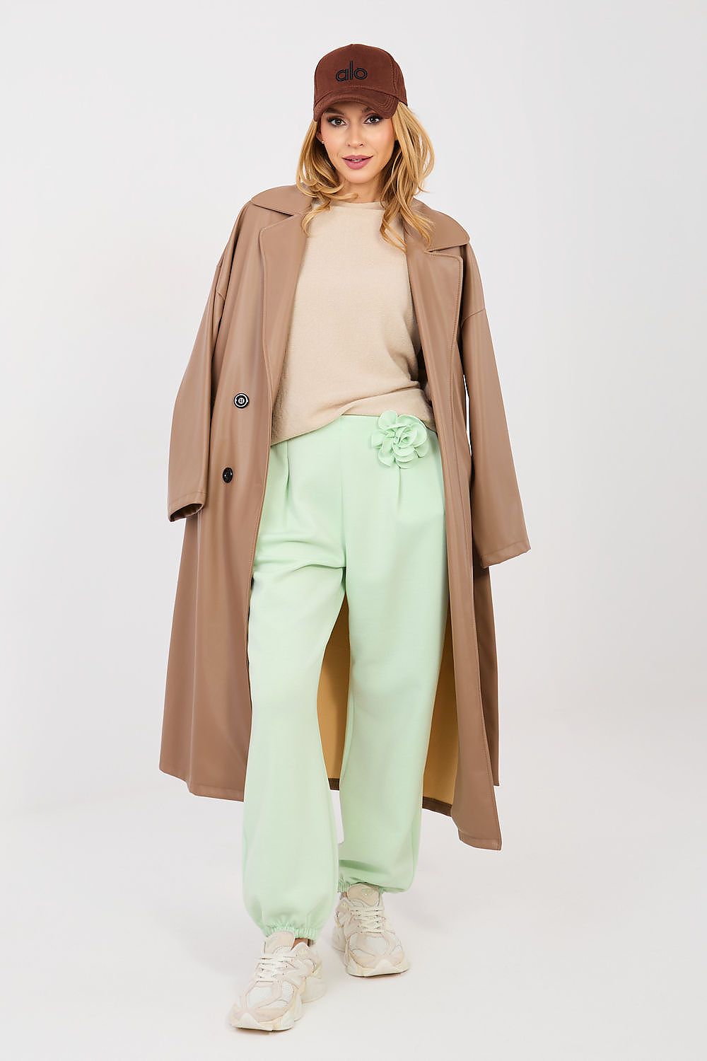 Woman wearing a beige coat, light green pants, and a brown cap on a white background