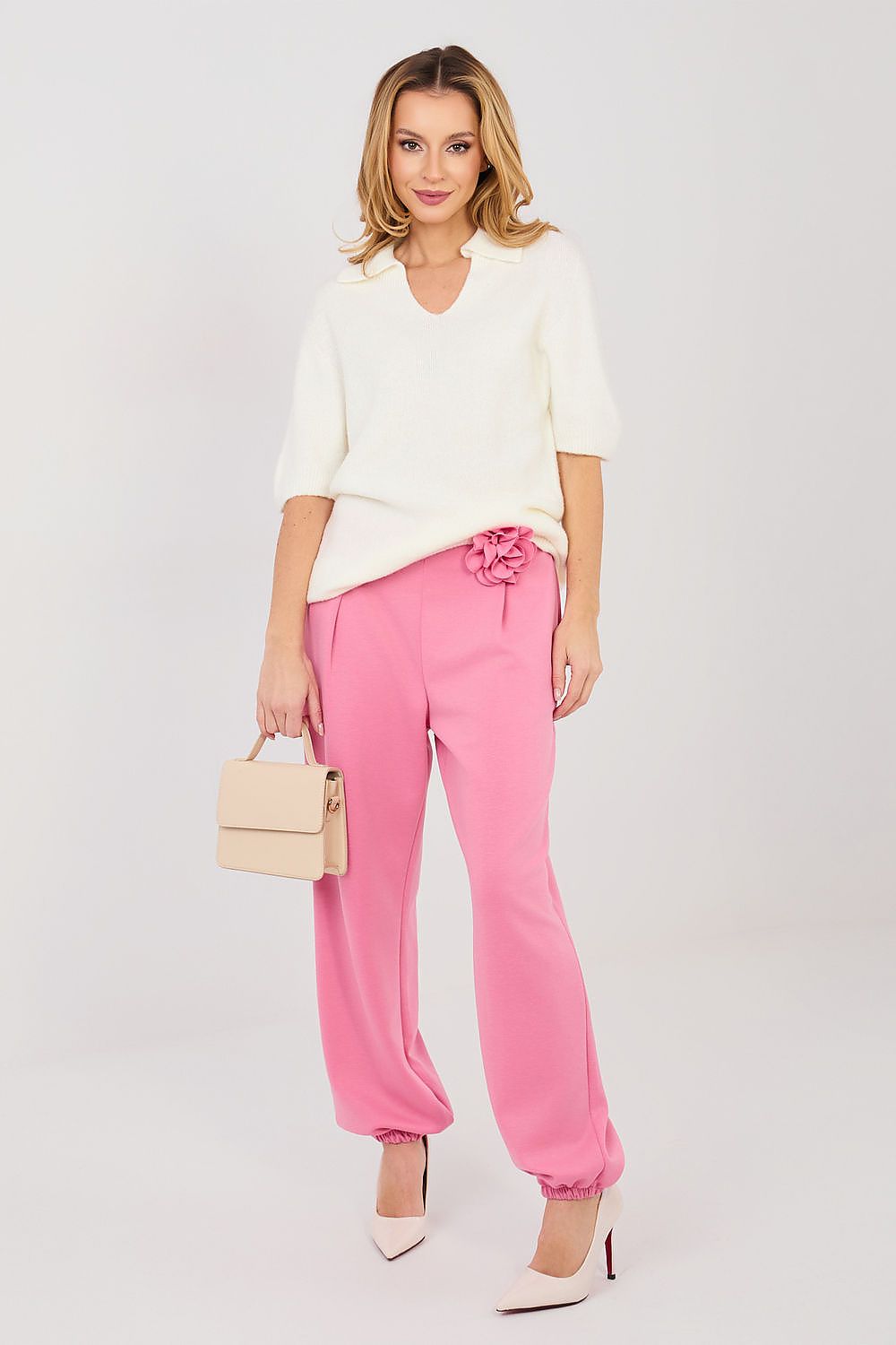 Woman wearing a white top and pink pants with a beige handbag on a light gray background