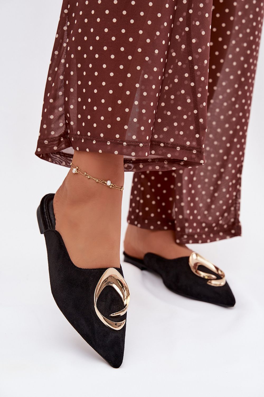 Black pointed-toe shoes with gold buckles worn with a brown polka dot outfit on a white background