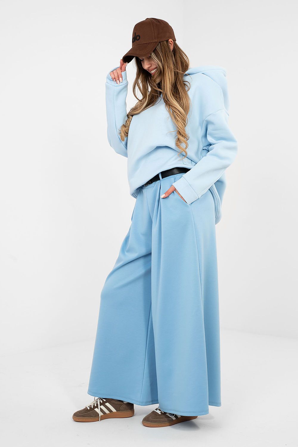 Person wearing a light blue outfit with wide-leg pants and a brown cap on a white background