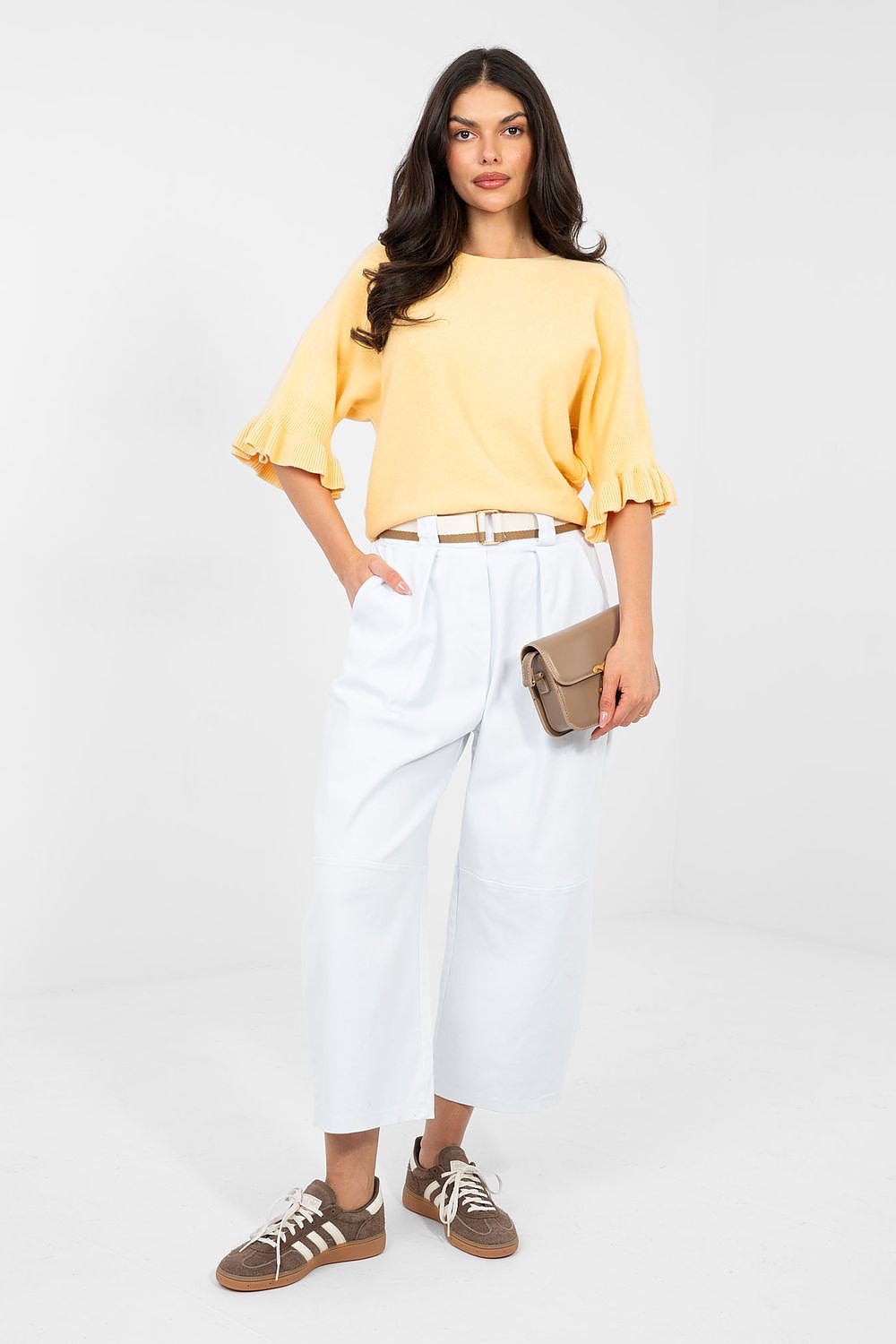 Woman wearing a yellow top and white pants with a beige clutch against a white background