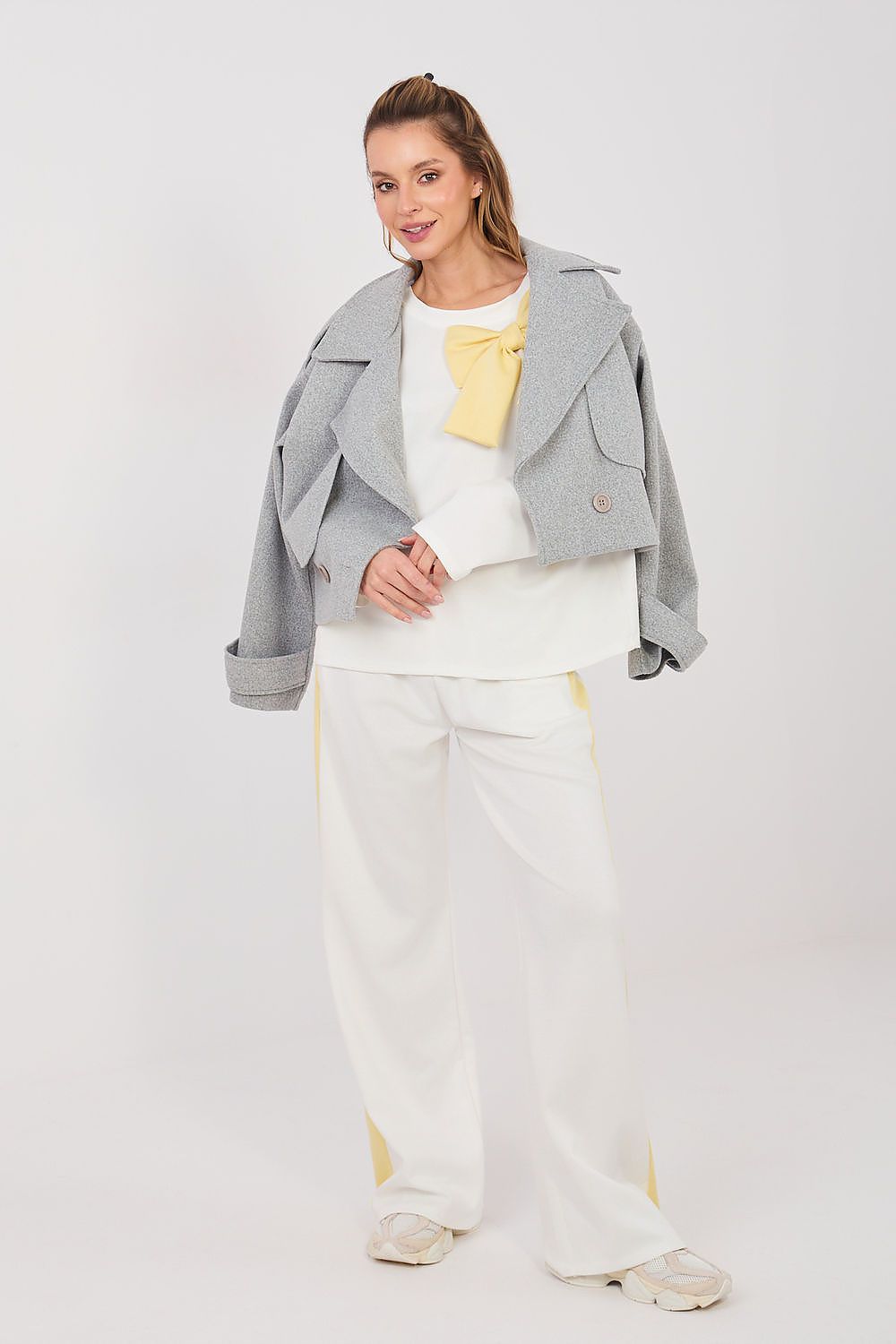 Woman wearing a gray jacket, white top, and white pants with yellow accents on a white background