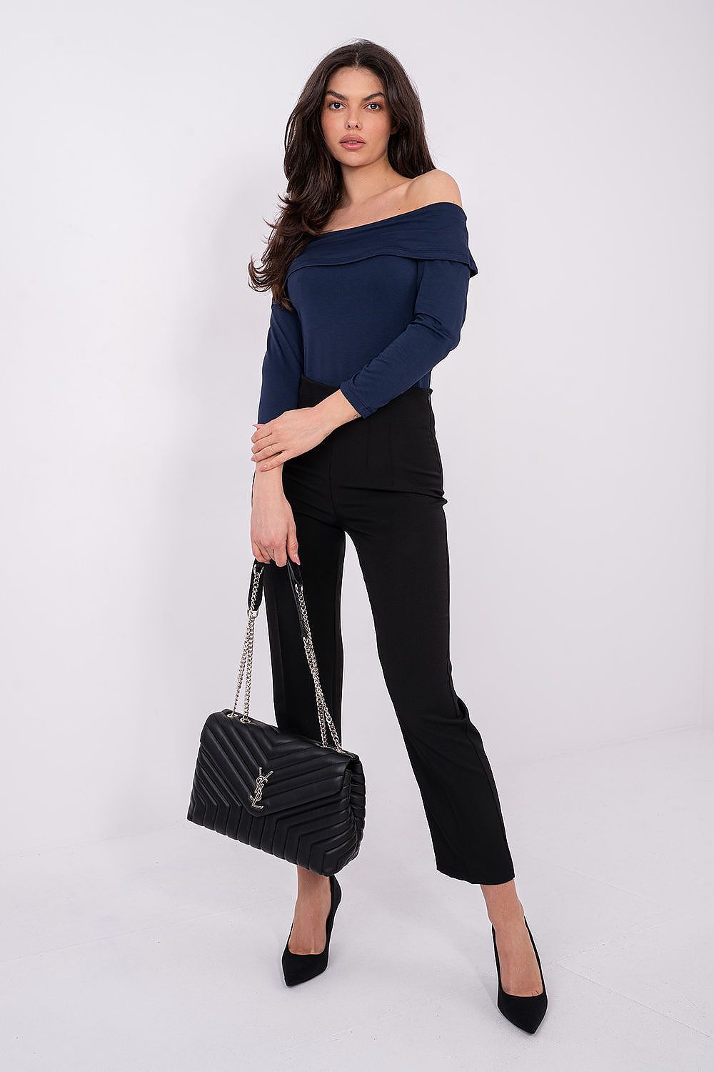 Woman wearing a navy off-shoulder top and black pants, holding a black handbag on a white background