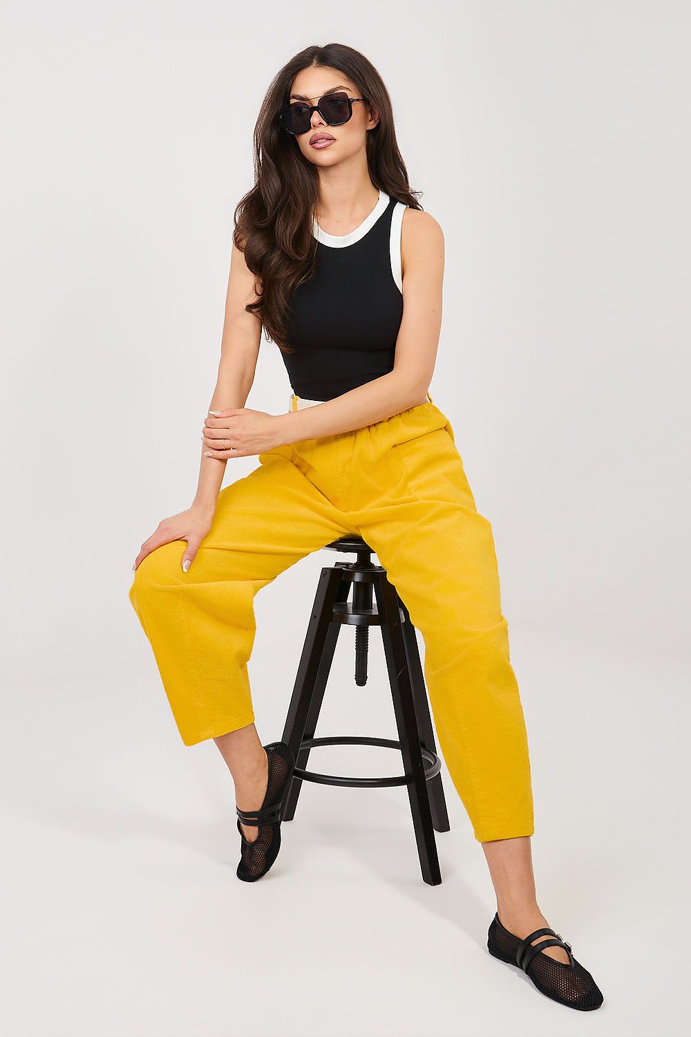 Woman sitting on a stool wearing yellow pants and black top against a white background