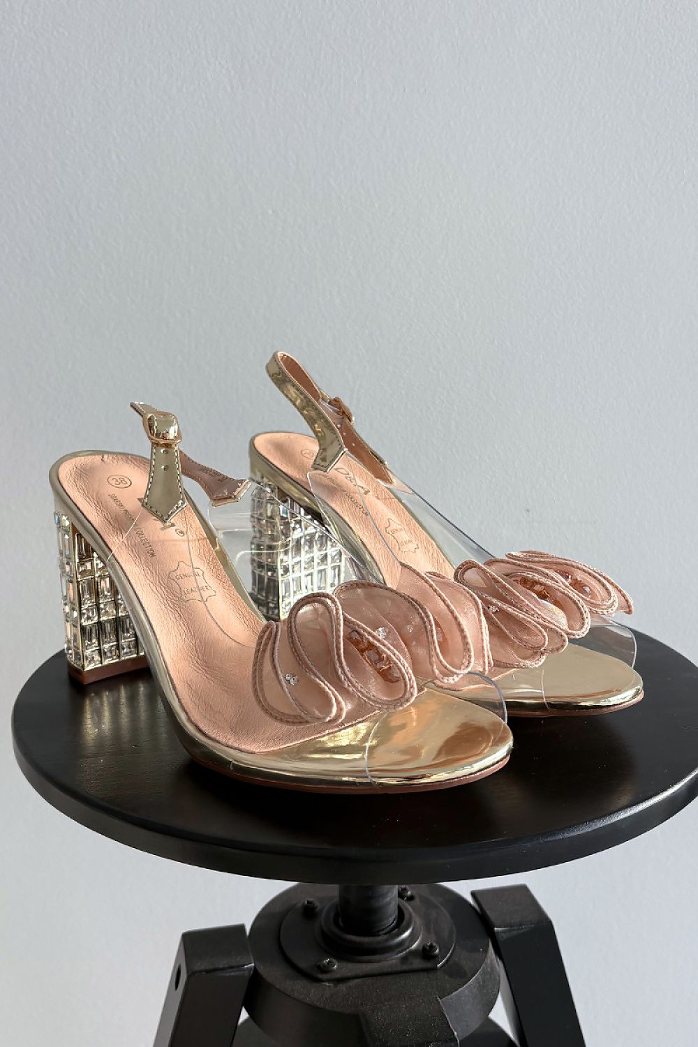 Pair of gold high-heeled shoes with clear straps on a black stand against a gray background