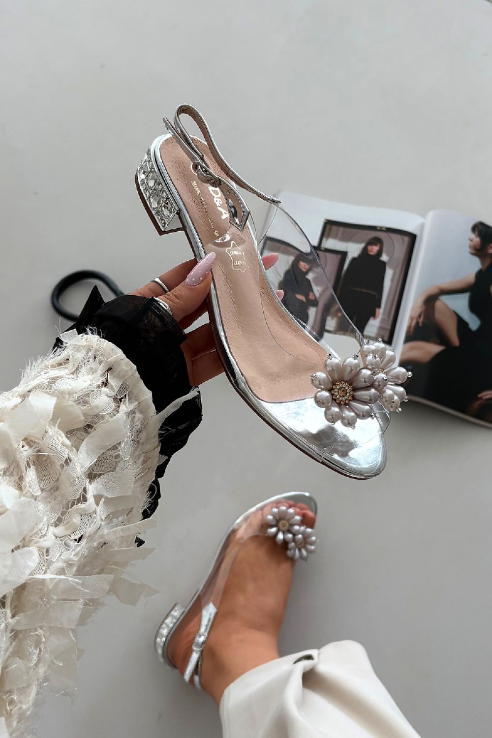 Silver high-heeled shoes with floral details worn by a person, on a light gray surface.