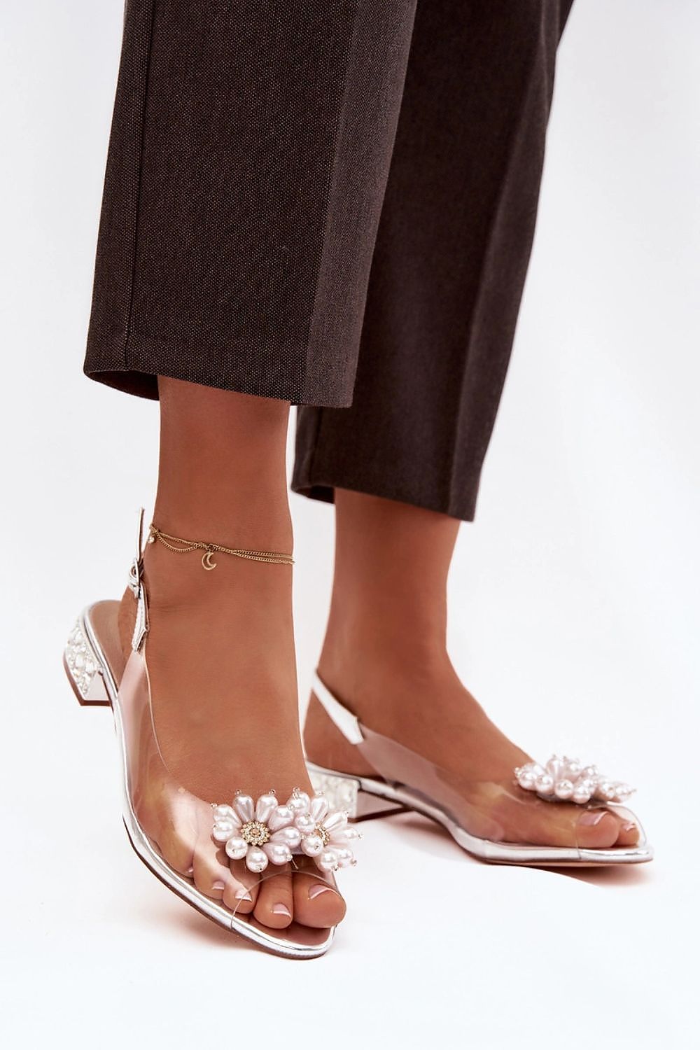 Clear high-heeled sandals with floral embellishments worn by a person in dark pants.