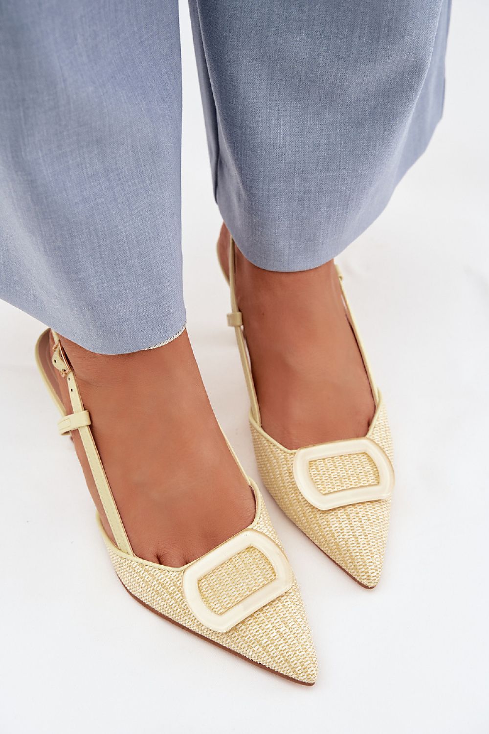 Beige pointed-toe flats with gold buckles worn with light blue pants on a white background
