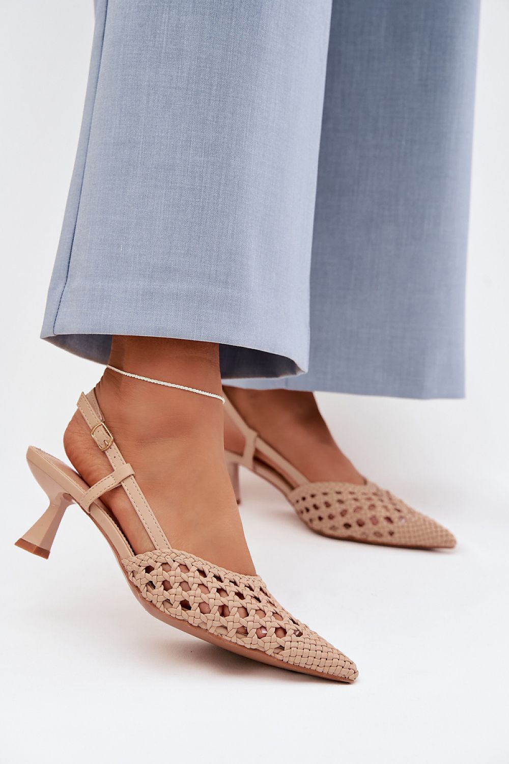 Beige high-heeled shoes with intricate detailing worn with light blue pants on a white background