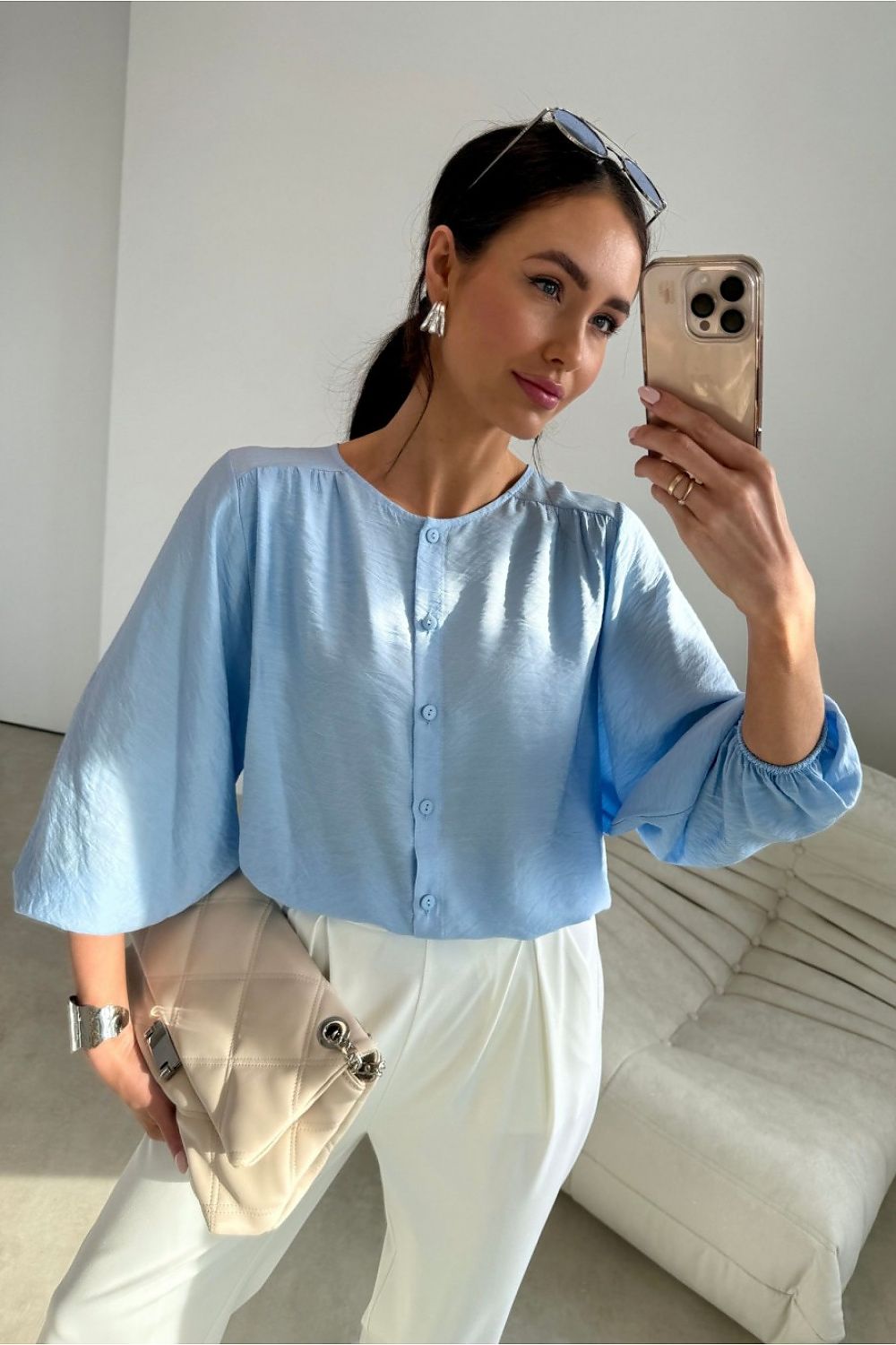 Woman taking a mirror selfie wearing a light blue shirt and white pants in a minimalistic room.