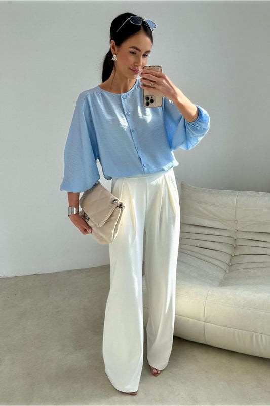 Woman taking a mirror selfie wearing a light blue shirt and white pants in a minimalistic room.