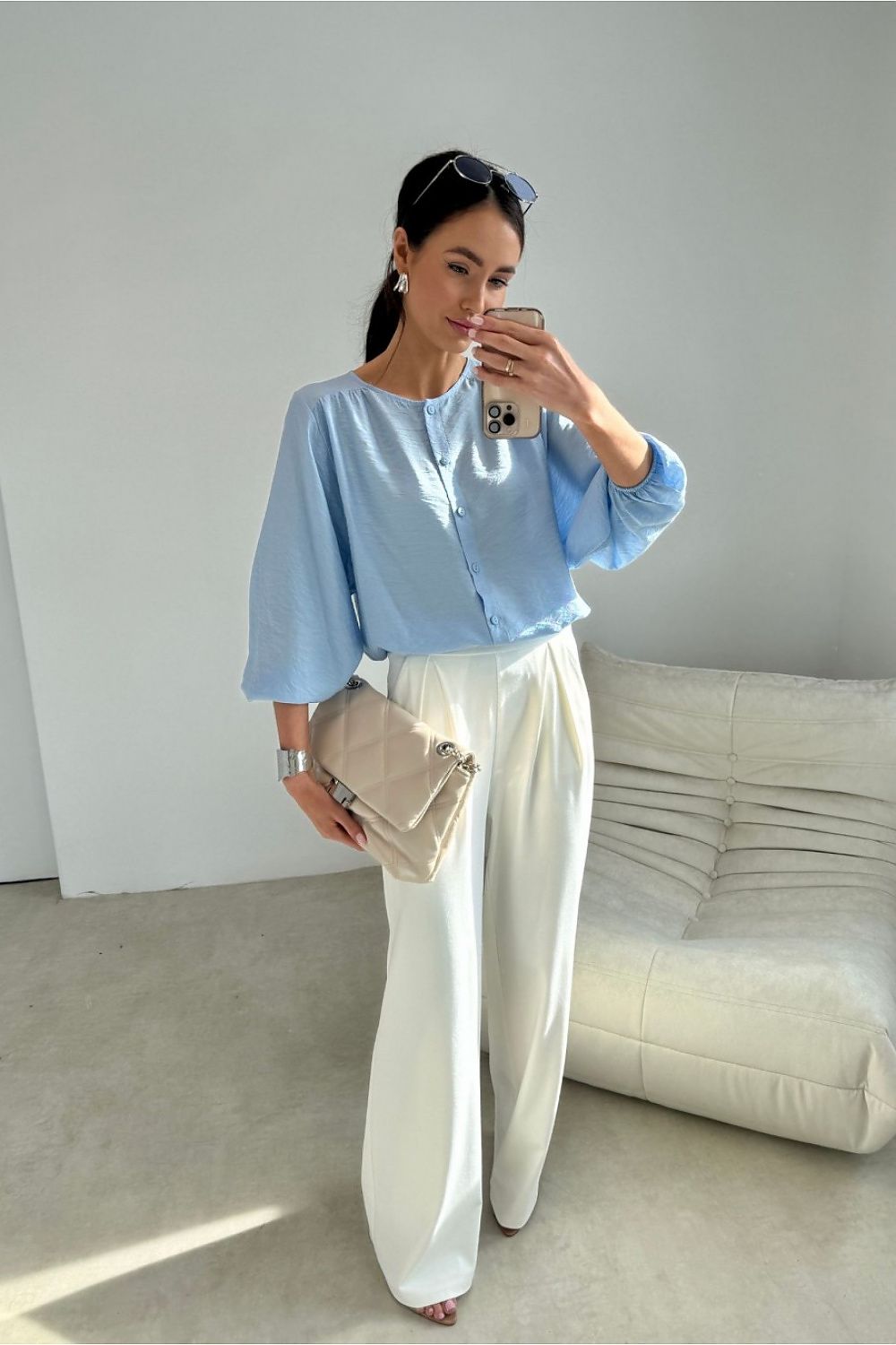 Woman taking a mirror selfie wearing a light blue blouse and white pants in a minimalistic room.