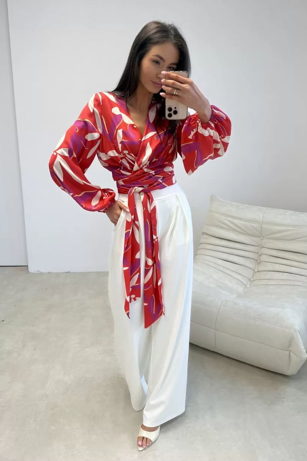 Woman taking a mirror selfie wearing a red and white patterned top and white pants in a minimalistic room.
