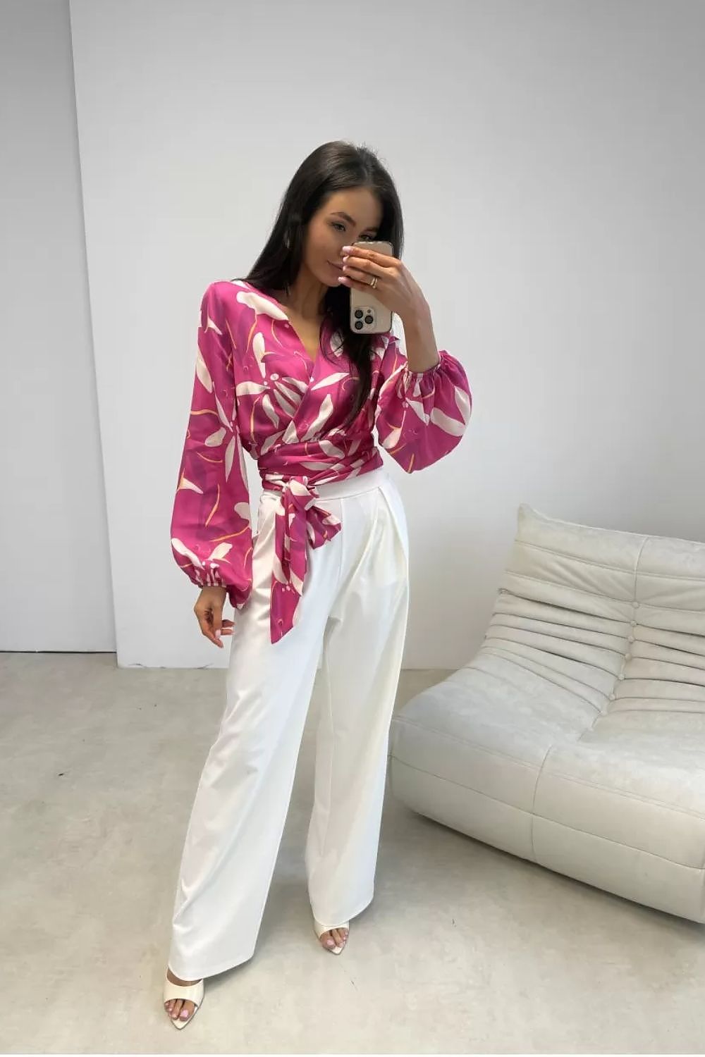 Woman wearing a pink and white patterned top with white pants in a minimalistic room.