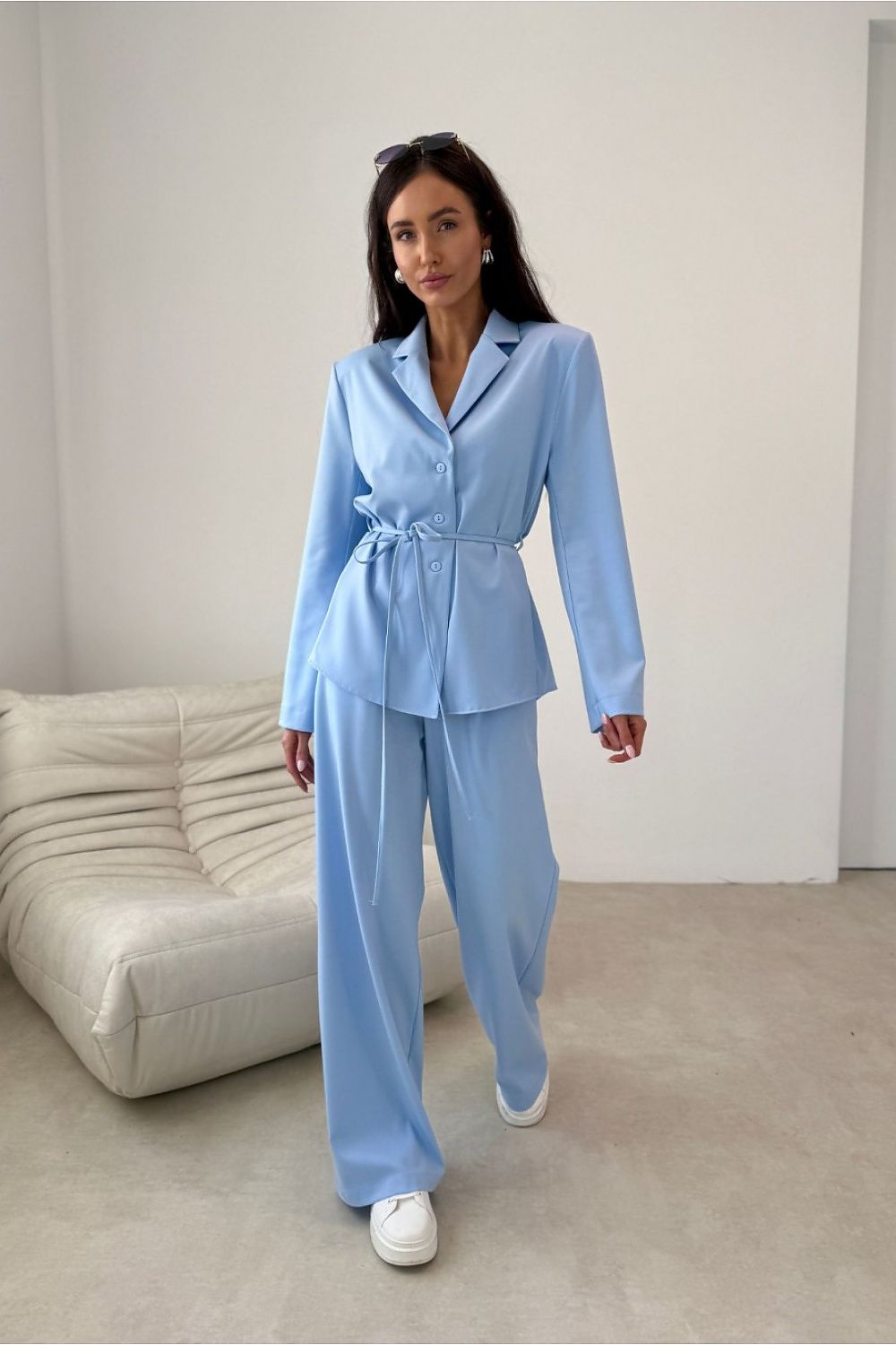 Woman wearing a light blue suit standing in a minimalistic room with a white sofa.