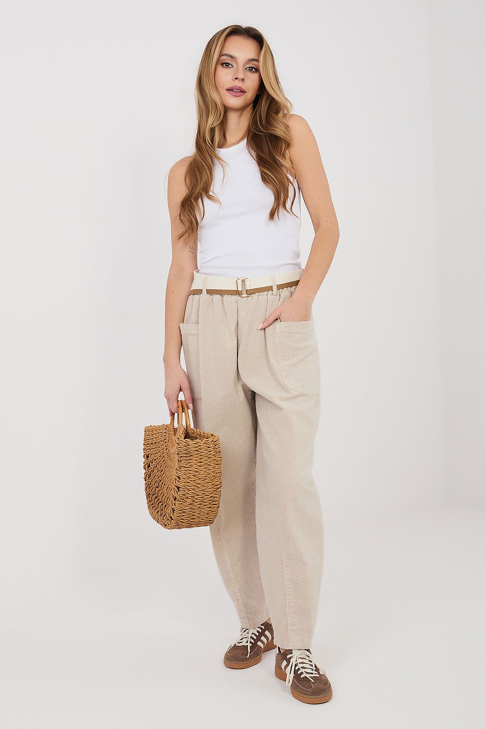 Woman wearing a white sleeveless top and beige pants, holding a woven bag on a white background