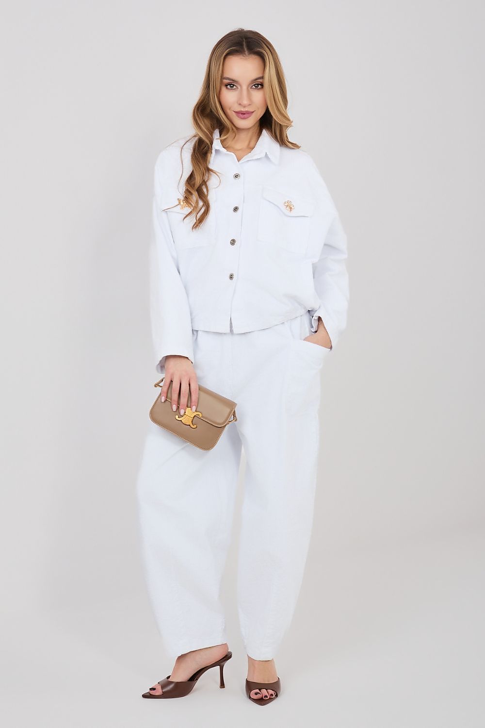 Woman in a white outfit holding a beige clutch against a plain background
