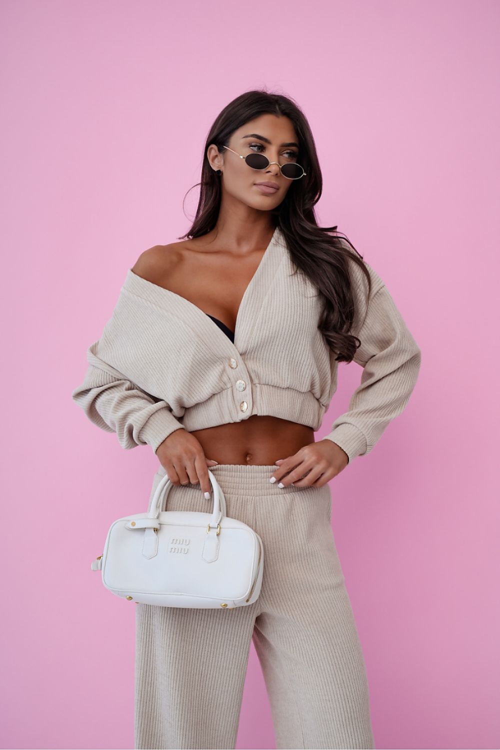 Woman in beige outfit with sunglasses and white handbag against a pink background