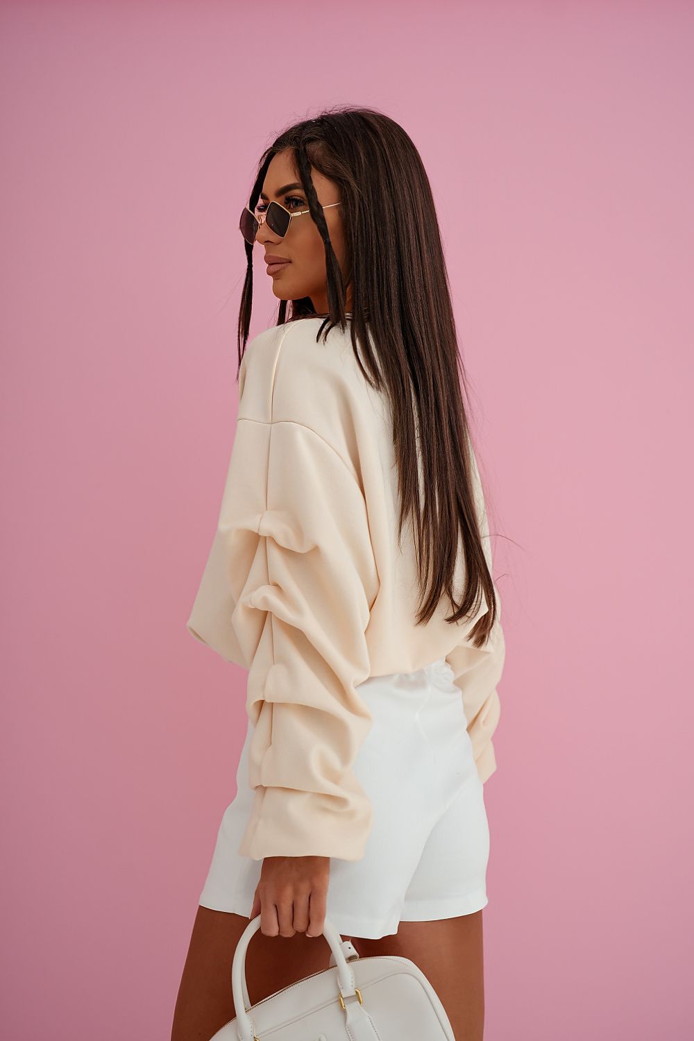 Woman wearing a beige sweatshirt and white shorts against a pink background