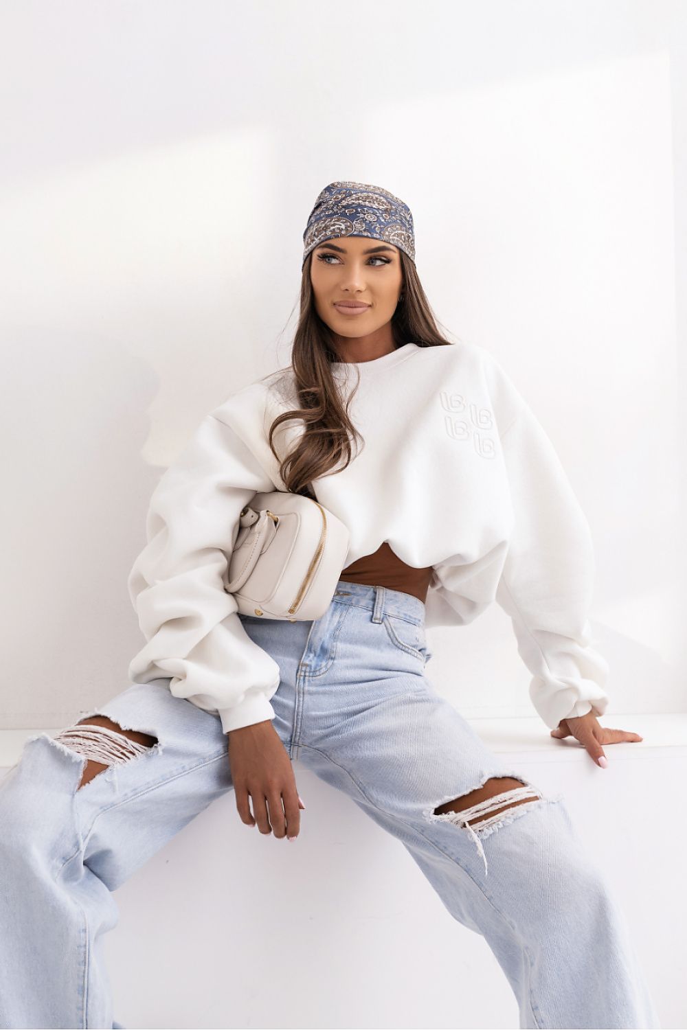 Woman wearing a white sweater and light blue jeans with a patterned headscarf, sitting on a white surface.