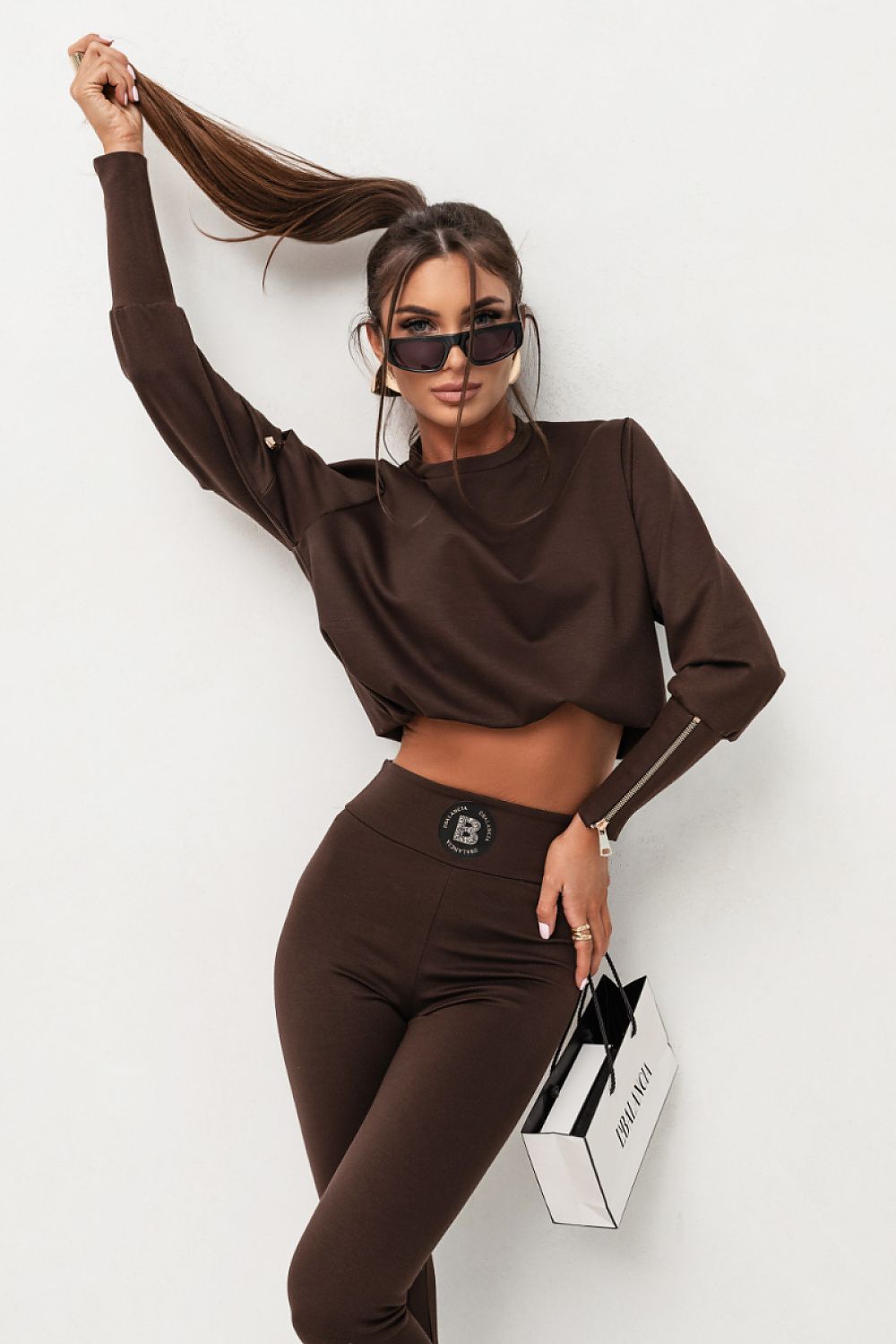 Woman in a brown outfit with sunglasses holding a black and white handbag on a white background