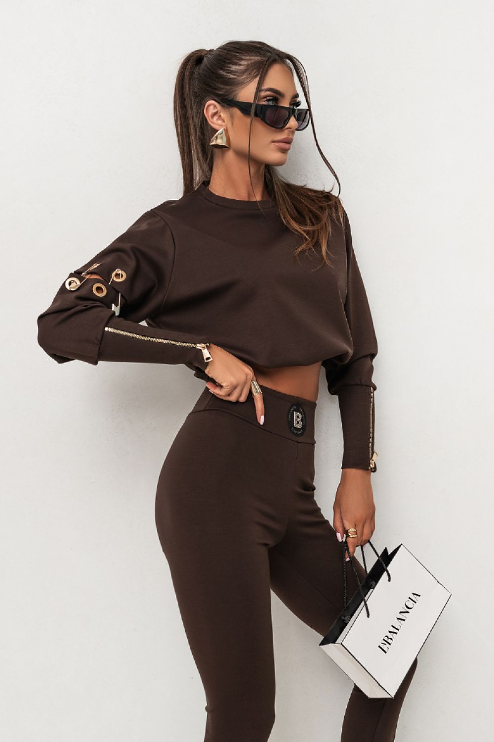 Woman in brown outfit holding a Prada bag against a white background