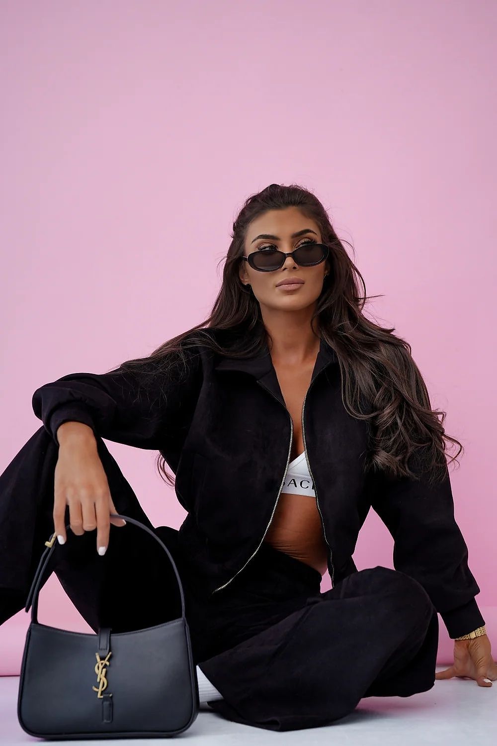Woman in black outfit with sunglasses and handbag against a pink background