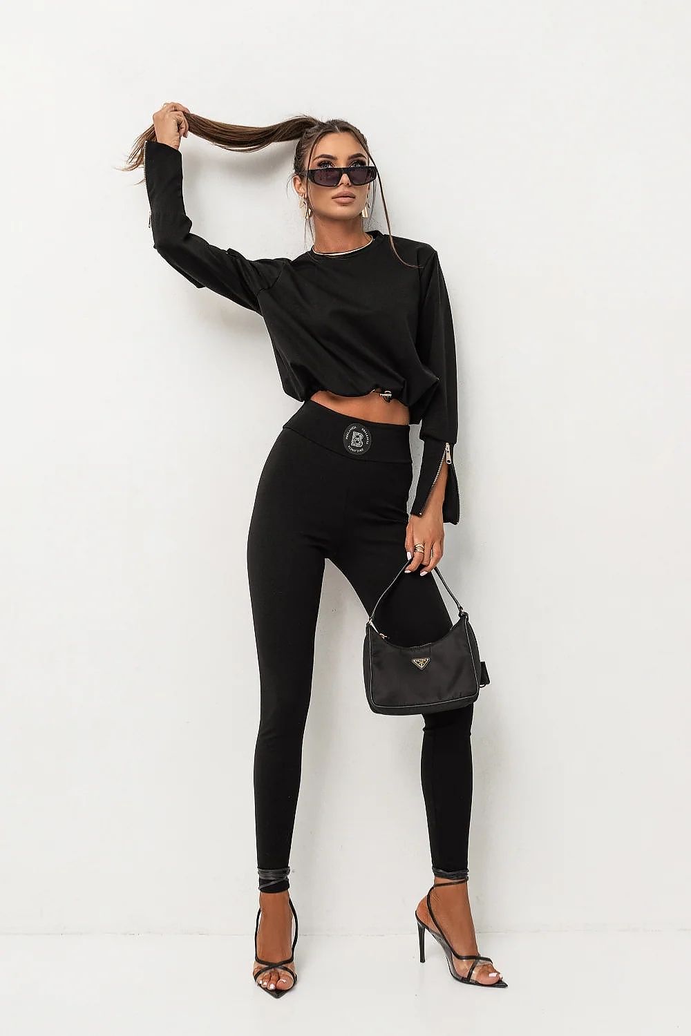Woman in black outfit with sunglasses and handbag against a white background