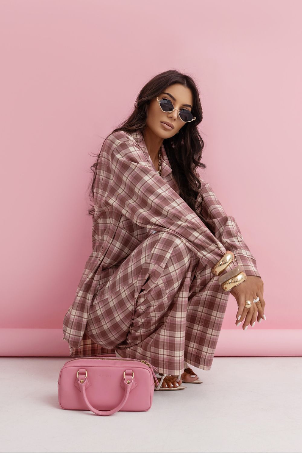 Woman in a plaid dress sitting on a pink couch with a pink handbag next to her.