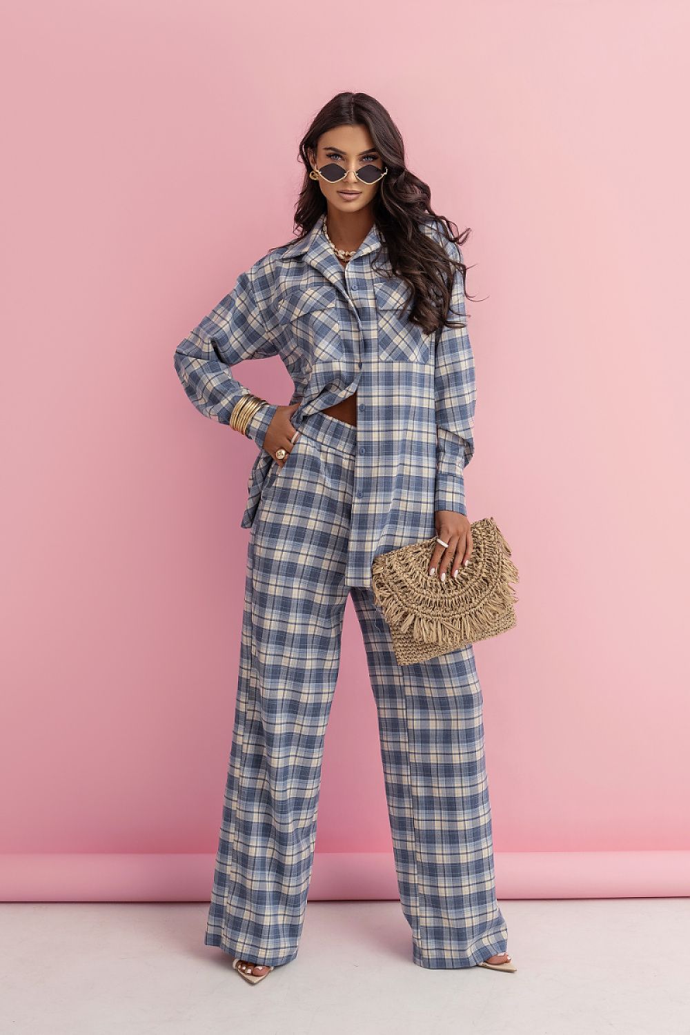 Woman in a blue plaid outfit holding a woven handbag against a pink background