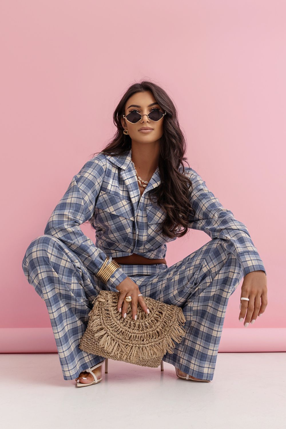 Woman in a blue plaid outfit holding a woven bag against a pink background