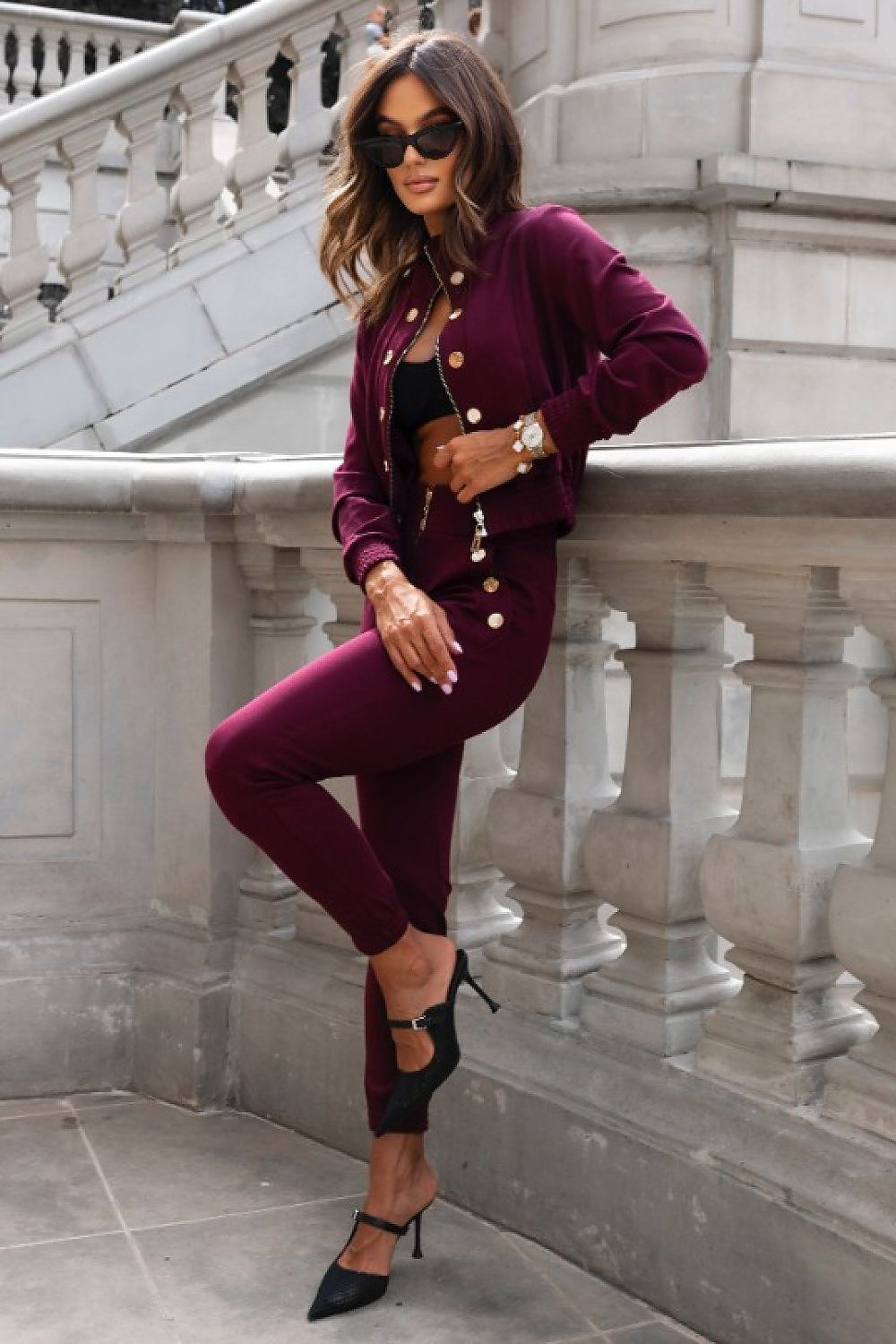 Woman in a burgundy outfit with gold buttons standing against a stone wall.