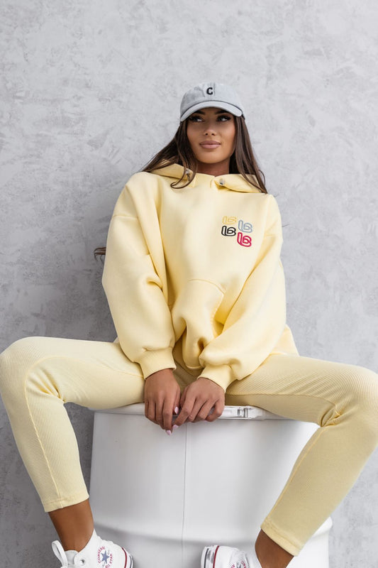Person wearing a yellow hoodie and pants sitting on a white stool against a gray background