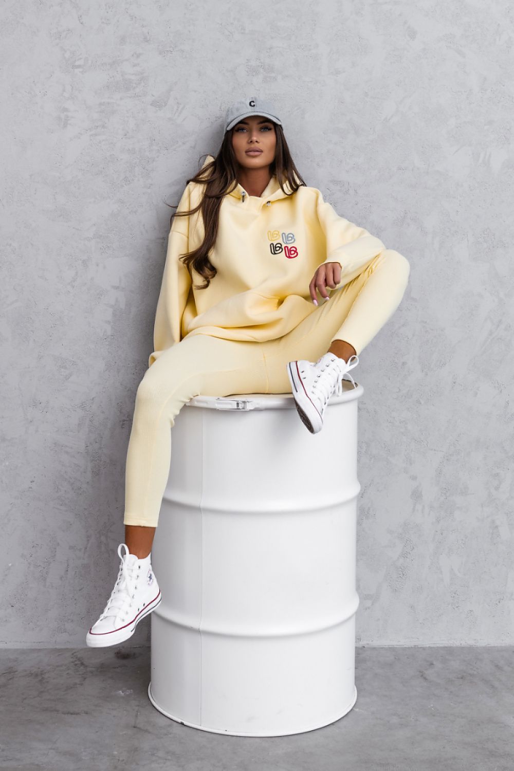Person wearing a yellow tracksuit sitting on a white barrel against a gray wall.
