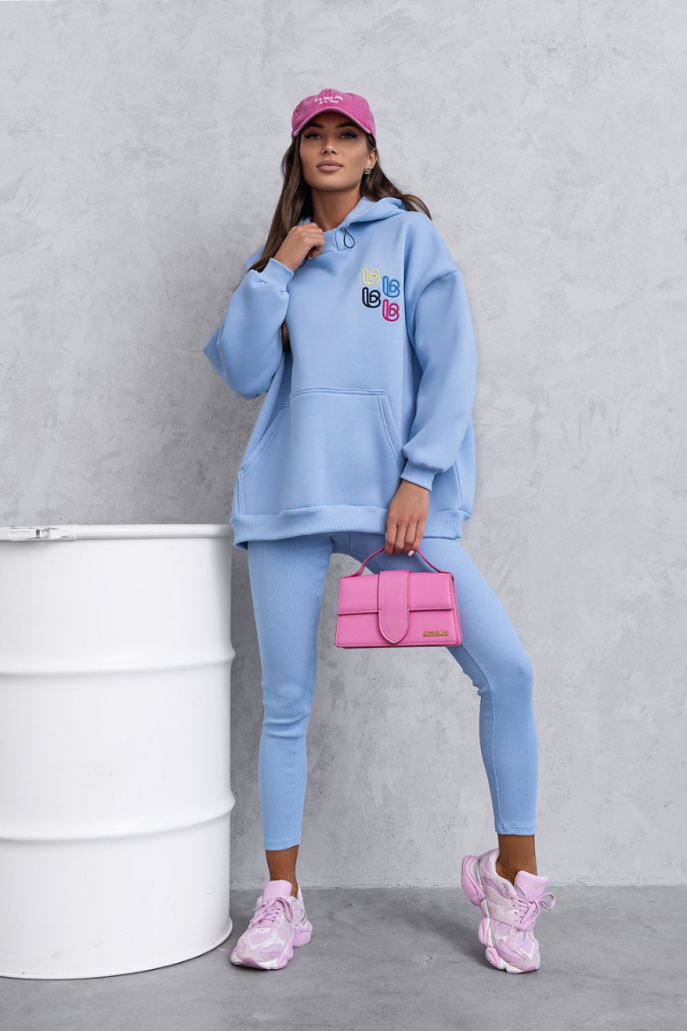Person wearing a blue tracksuit with a pink cap and bag, standing against a gray background.