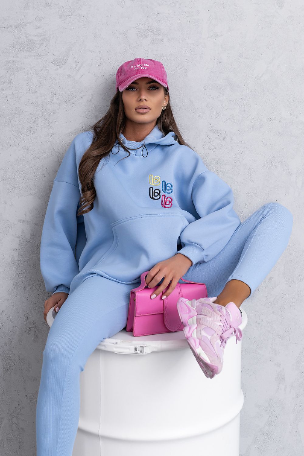 Person wearing a light blue hoodie and pants with colorful logo, sitting on a white stool against a gray background.