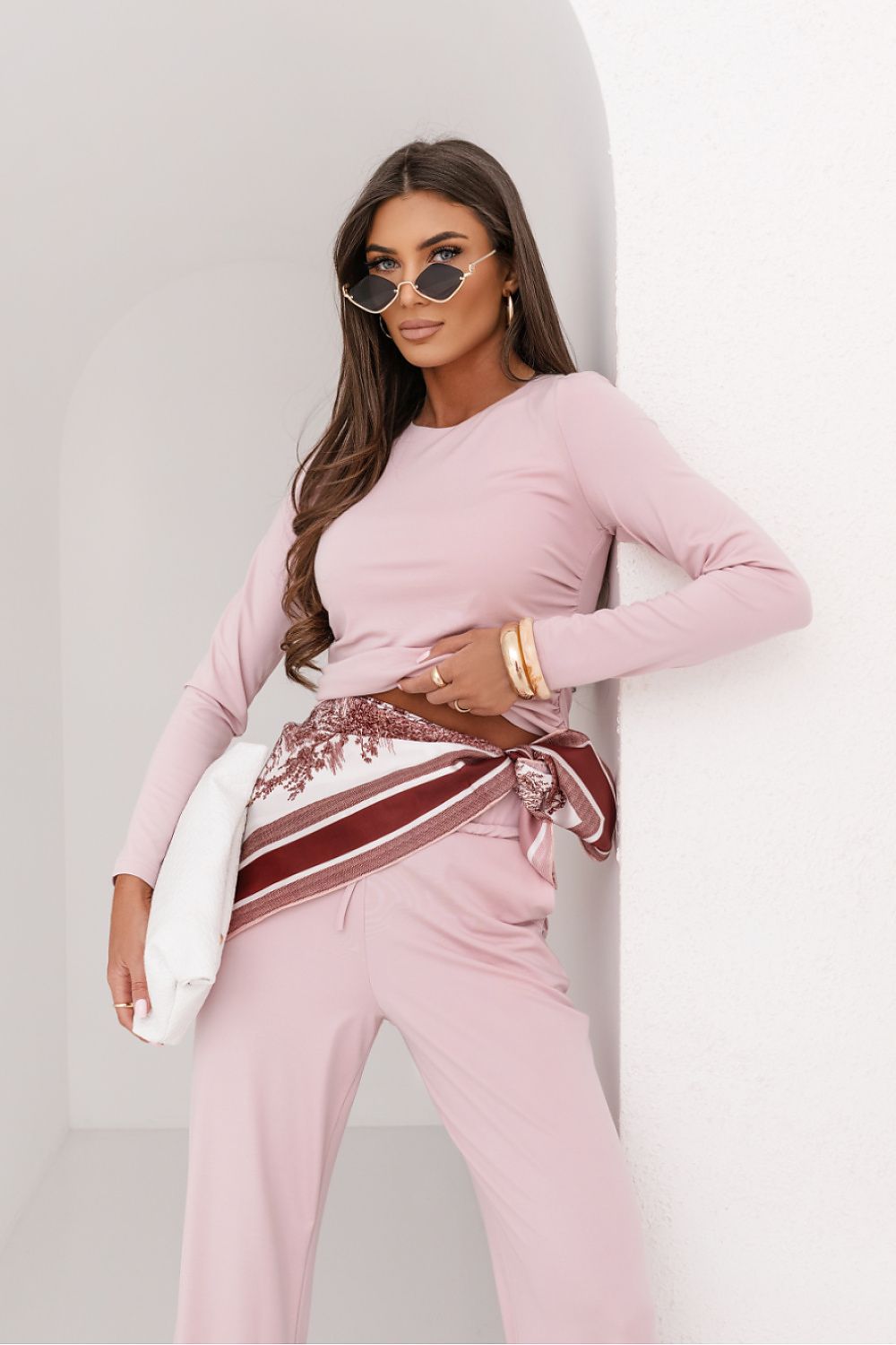 Woman wearing a pink outfit with a belt and sunglasses against a light background