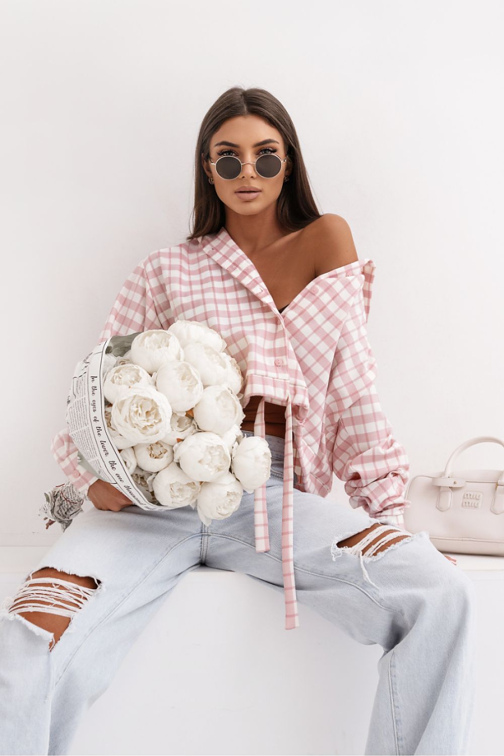 Woman holding a bouquet of white flowers wearing a pink checkered shirt and light blue jeans.